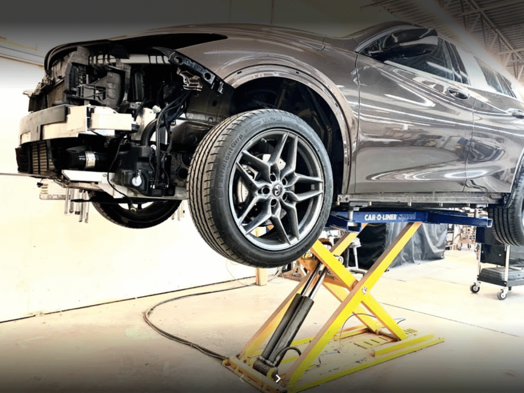 A grey sedan with front bumper damage is lifted on hydraulics in the middle of an active garage.
