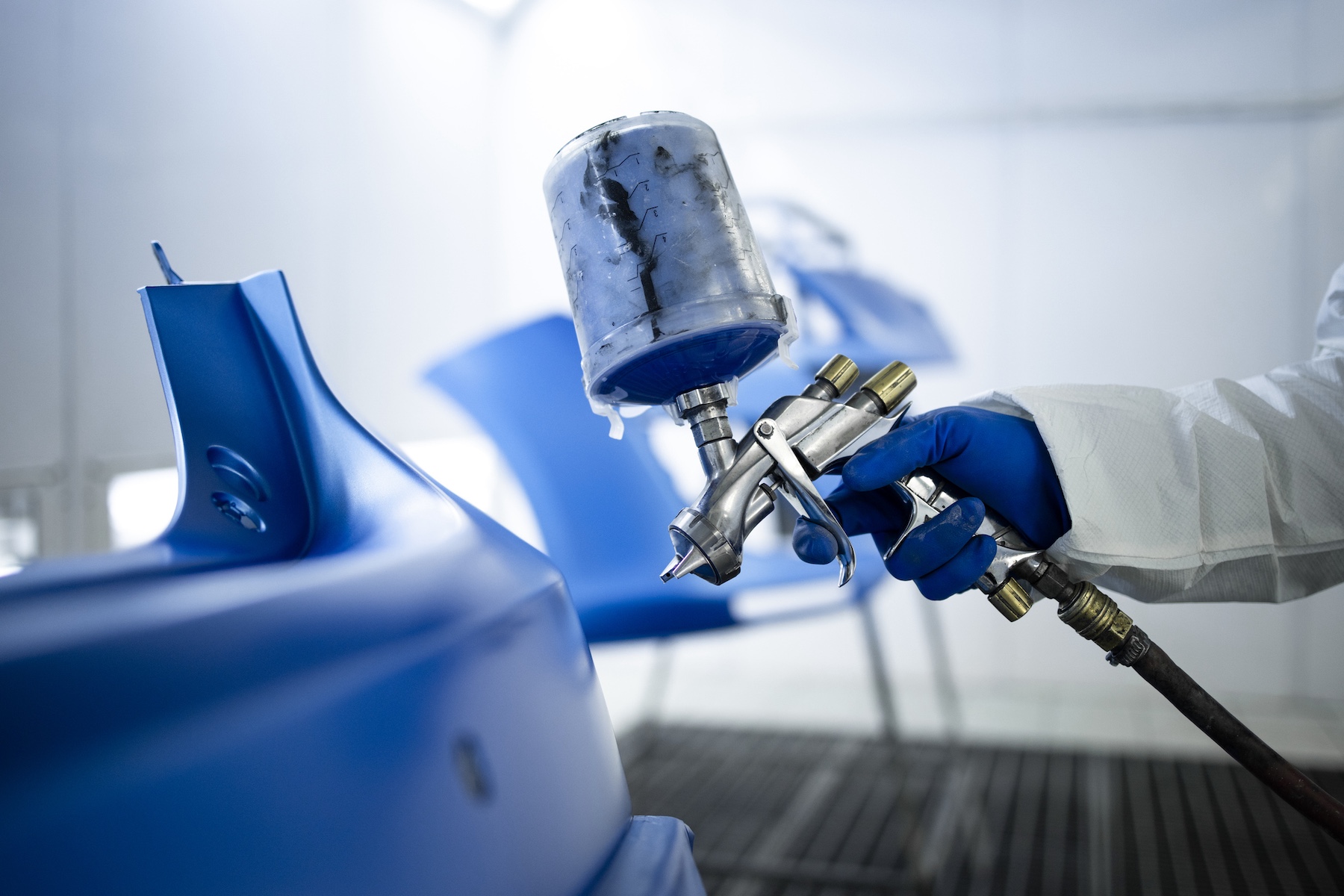 Car painter in protective clothes and mask painting automobile bumper with metallic paint and varnish in chamber workshop.
