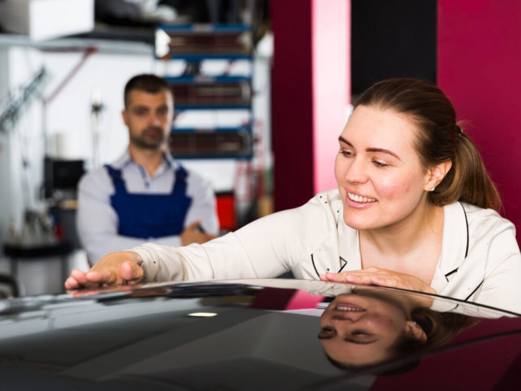 A woman with brown hair and a poytail wearing a white jacket smiles as she assesses her newly repaired vehicle, while a technician looks on in the background.